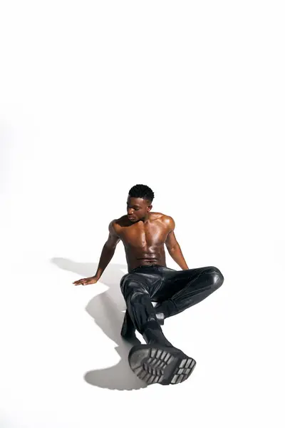 A young African American man sits confidently on the floor, bare chested, wearing stylish black pants. — Stock Photo