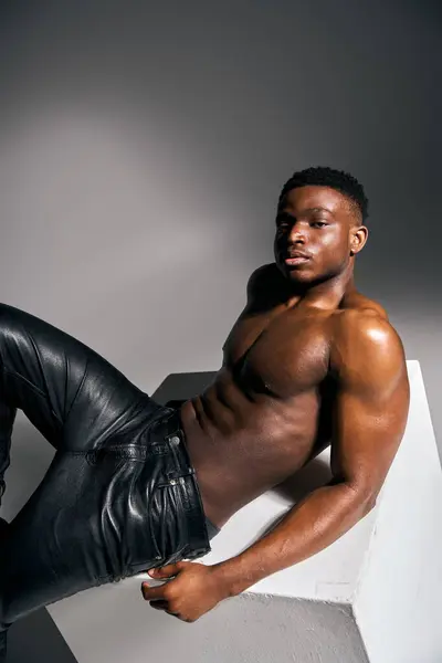 A young and attractive African American man showcases his physique while striking a bold pose, dressed in edgy black leather pants. The minimalist setting highlights his confidence. — Stock Photo