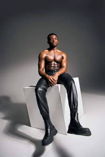 Um jovem e atraente afro-americano posa topless em um pedestal branco, exalando confiança e força. Ele usa calças pretas elegantes e botas ousadas, cercado por um pano de fundo minimalista. — Fotografia de Stock