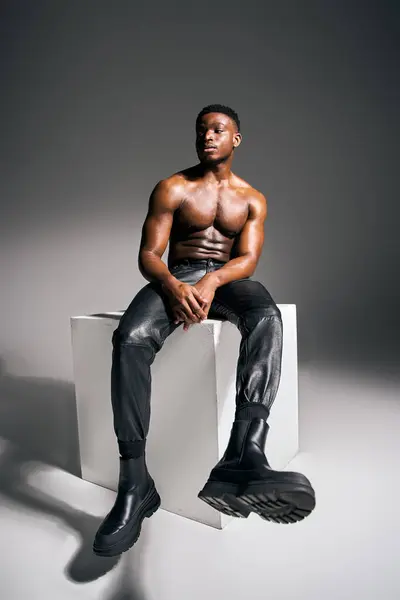Um jovem e atraente afro-americano posa confiantemente sem camisa em um estúdio contemporâneo. Ele se senta em um cubo, vestindo calças de couro elegantes e botas, incorporando força e carisma. — Fotografia de Stock