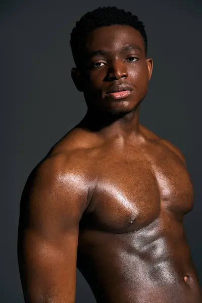 A young African American man stands confidently, showcasing his sculpted torso against a dark backdrop. His expression is captivating, reflecting strength and charisma in this striking portrait. — Stock Photo