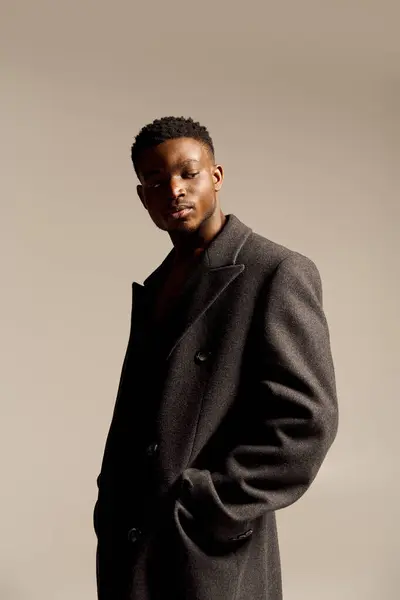 A young handsome African American man stands tall, radiating confidence while wearing a chic coat. The warm backdrop enhances his striking features as he strikes a relaxed pose, showcasing elegance. — Stock Photo