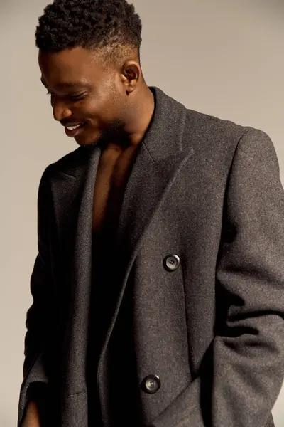 Un giovane uomo afroamericano bello si alza con fiducia, mostrando il suo fisico tonico mentre indossa un cappotto alla moda. È al chiuso, trasuda fascino e carisma. — Foto stock