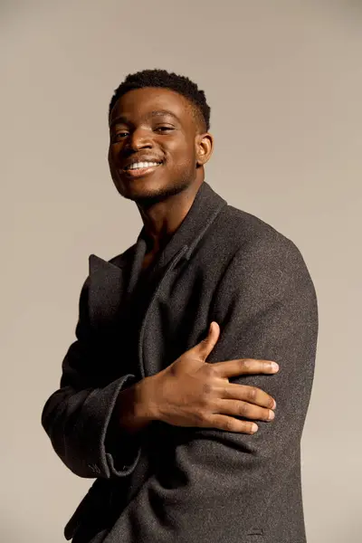 Un jeune homme afro-américain séduisant montre son sourire confiant tout en posant avec une position forte. Il porte un manteau chic, capturant un moment de charme et de sophistication dans un cadre minimaliste. — Photo de stock