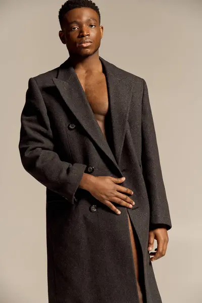 A young, handsome African American man poses confidently in a sleek coat, showcasing his charm. The neutral background enhances his striking features and adds an artistic flair to the moment. — Stock Photo