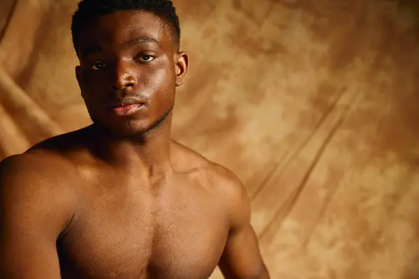 A young and attractive African American man showcases his physique with a confident expression. The warm background enhances his striking features, creating an inviting atmosphere. — Stock Photo