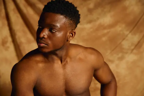 A confident young African American man poses topless against a warm toned backdrop, showcasing his sculpted physique. His keen expression conveys a sense of allure and charm. — Stock Photo