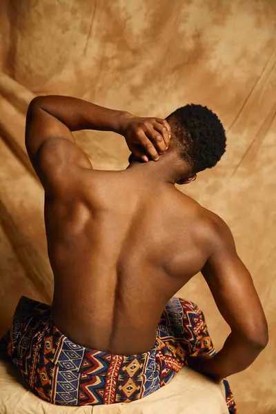 A young handsome african american man poses topless with grace, showcasing muscular form while sitting elegantly against a textured backdrop, highlighting creativity and confidence in his body. — Stock Photo
