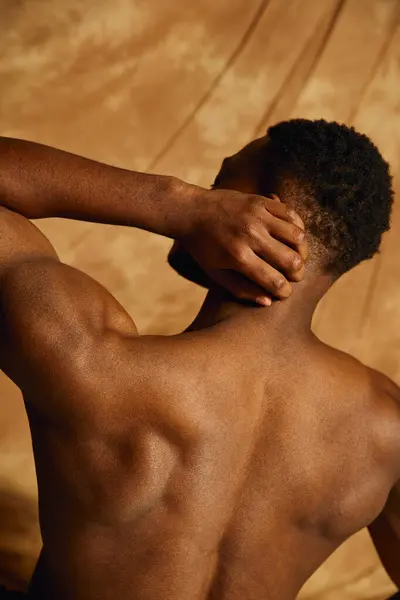 An attractive young African American man poses topless, revealing a toned physique while playfully touching his neck. The warm toned backdrop adds an artistic touch, enhancing the mood. — Stock Photo
