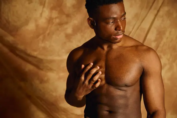 A young, handsome African American man poses effortlessly without a shirt, showcasing his strong physique. The warm backdrop enhances his features while he gazes thoughtfully to the side. — Stock Photo