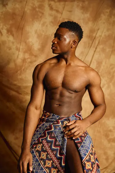 A young and fit African American man stands topless, showcasing his physique. Draped in colorful fabric, he gazes thoughtfully to the side, immersed in the creative atmosphere of the studio. — Stock Photo