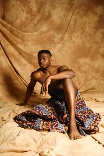 A young and striking African American man poses confidently without a shirt, draped in a patterned fabric. The warm backdrop enhances his features, emphasizing his relaxed and artistic vibe. — Stock Photo