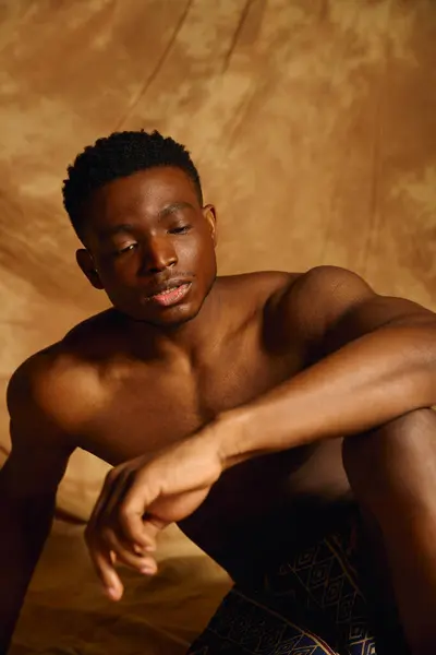 A young handsome African American man sits gracefully, showcasing his striking physique and relaxed demeanor. The warm backdrop enhances the intimate mood, highlighting his natural charisma. — Stock Photo