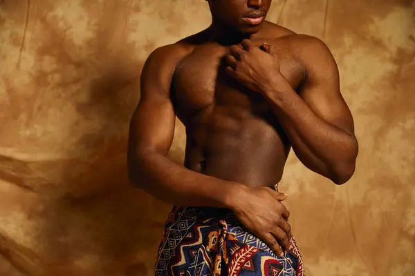 A young African American man stands confidently, posing topless. He highlights his well defined physique while draped elegantly in patterned fabric, against a textured background. — Stock Photo