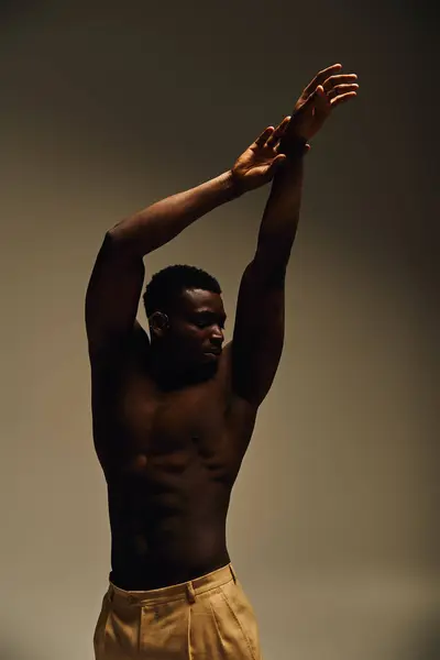 A young handsome African American man strikes a pose, confidently showcasing his physique. He stands tall, arms raised, embodying strength and artistry in a subdued indoor atmosphere. — Stock Photo