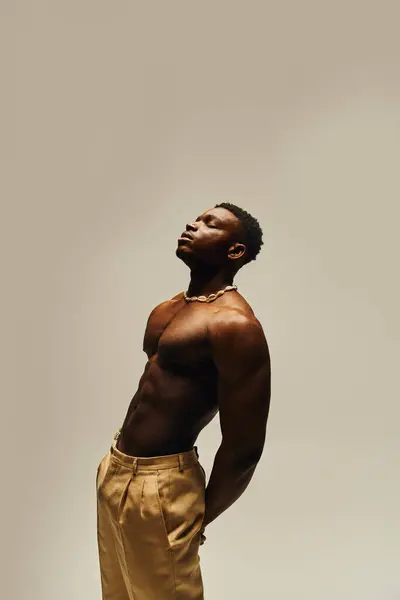 Striking a pose, a young handsome African American man stands topless, exuding confidence and charm. He combines fashion forward trousers with a relaxed posture, drawing attention to his physique. — Stock Photo