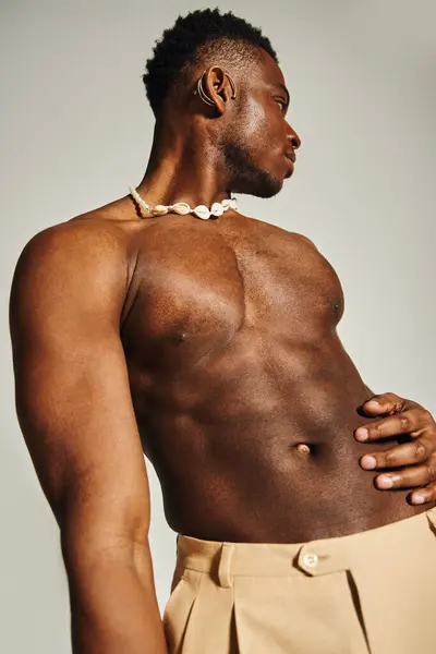 A young, handsome African American man stands confidently, showcasing his fit physique. He poses stylishly with a playful expression, wearing fashionable trousers and a unique necklace. — Stock Photo