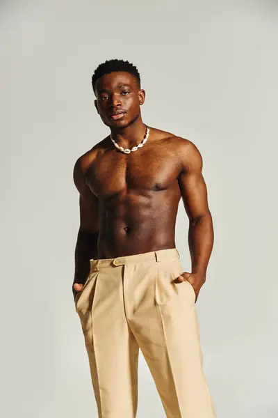 A young handsome African American man poses confidently without a shirt, wearing stylish beige trousers and a necklace. The neutral background enhances his strong physique and striking presence. — Stock Photo