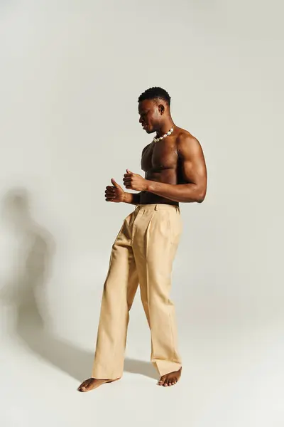 Un giovane uomo afroamericano bello si alza con fiducia, mostrando il suo fisico tonico. Vestito con semplici pantaloni beige, trasuda carisma su uno sfondo pulito, coinvolgendo lo spettatore con il suo portamento.. — Foto stock