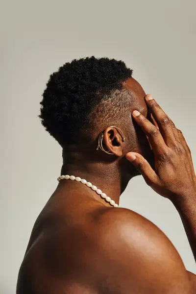 Un bel giovane afroamericano in topless posa con stile, coprendo strategicamente il suo viso con una mano. I suoi capelli corti ricci e una semplice collana esaltano il suo aspetto sorprendente. — Foto stock