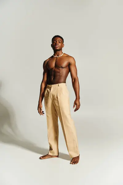 A young handsome African American man stands confidently, showcasing a strong physique without a shirt. He wears light colored trousers and a chain, creating a modern, stylish appearance. — Stock Photo