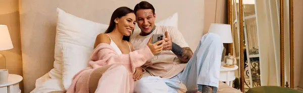 Young couple cuddles together on a bed, sharing laughs while looking at a phone screen. — Stock Photo