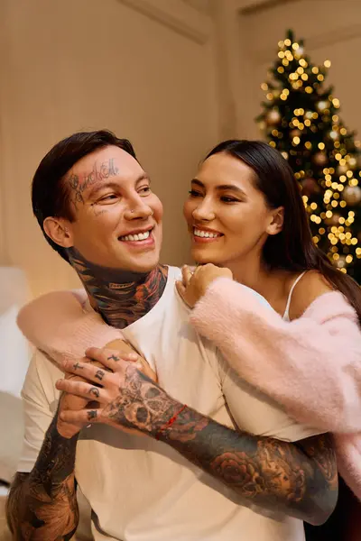 Two young lovers embrace warmly while smiling by the festive lights of a holiday tree. — Stock Photo