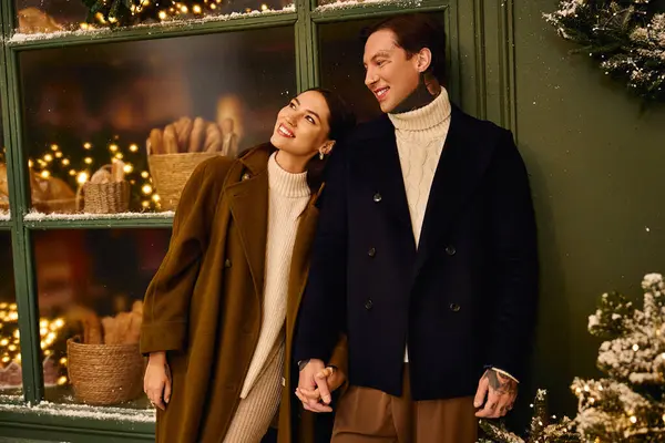 Pareja cariñosa sonríe cálidamente mientras se toma de la mano cerca de la pantalla de la ventana festiva con luces brillantes - foto de stock