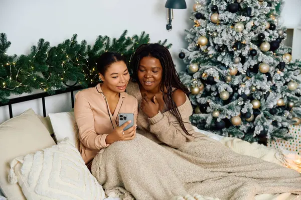 Two women relax with a warm blanket, browsing a smartphone by holiday decor. — Stock Photo
