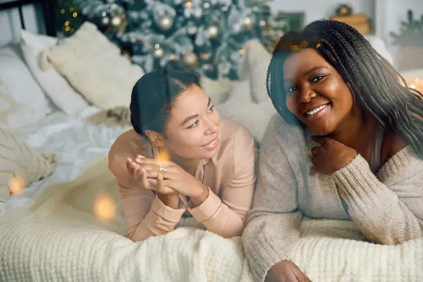 Two women relax together on a bed, surrounded by warm blankets and holiday decor. — Stock Photo