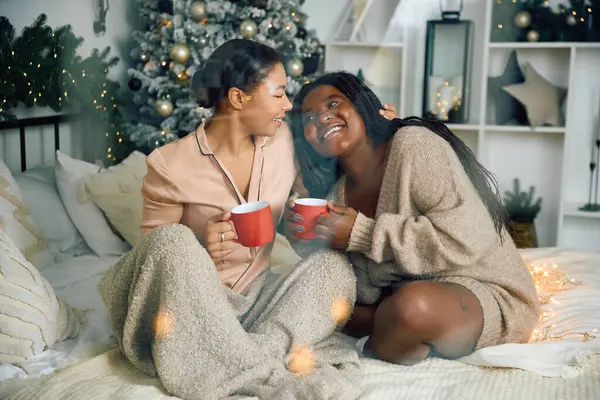 Deux femmes s'assoient ensemble sur un lit, souriant et tenant des tasses rouges dans un cadre hivernal festif. — Photo de stock