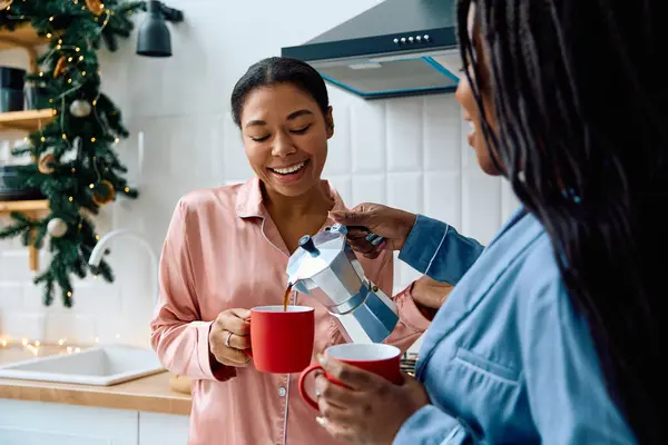 Dos mujeres sonríen mientras una vierte café en una taza roja, llenando su cocina con calor. - foto de stock