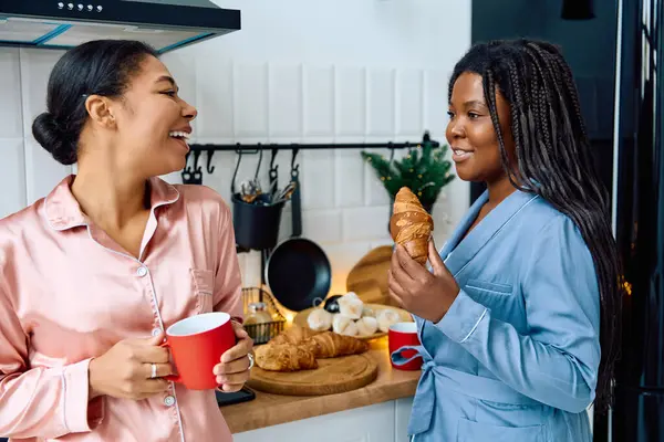 Two women enjoy breakfast together, laughing and sharing pastries in a warm kitchen setting. — Stock Photo