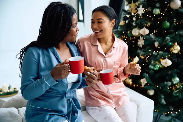 Two women in cozy pajamas enjoy hot drinks and pastries by a festive Christmas tree. — Stock Photo