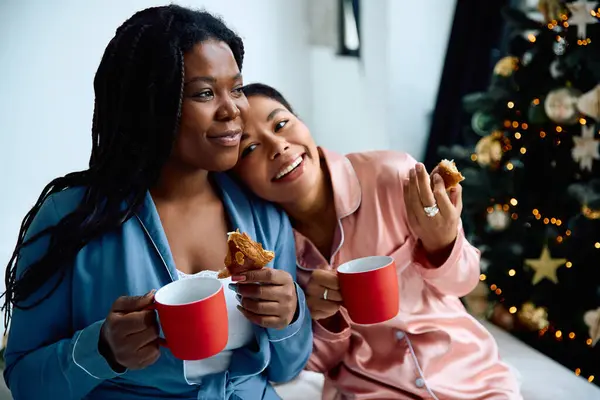 Two women relax together in their pajamas, enjoying cookies and warm drinks by a Christmas tree. — Stock Photo