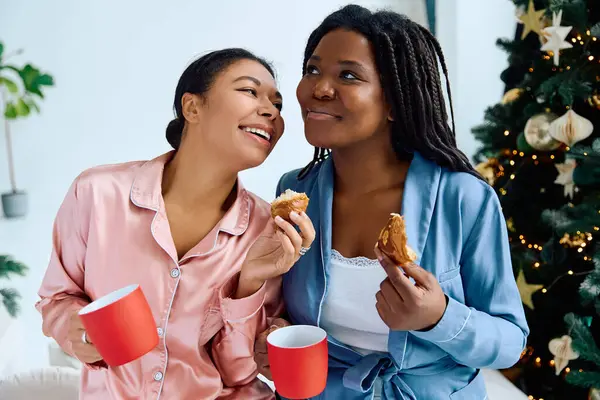 Two women share joyful moments while savoring coffee and pastries by the Christmas tree. — Stock Photo