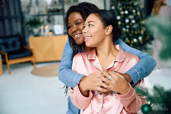 Two women smile warmly, embracing in their festive, holiday decorated living room. — Stock Photo