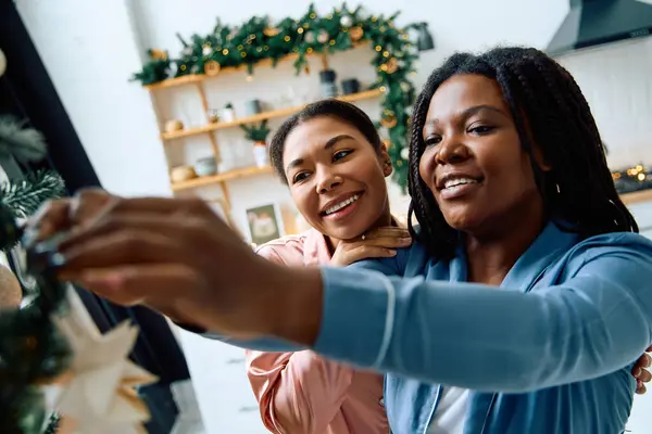 Two women happily decorate a Christmas tree with ornaments, sharing smiles and warmth. — Stock Photo