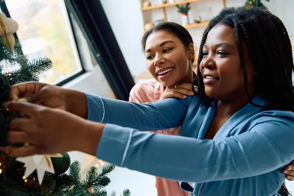 Two women joyfully decorate a Christmas tree together, embracing love and holiday cheer. — Stock Photo