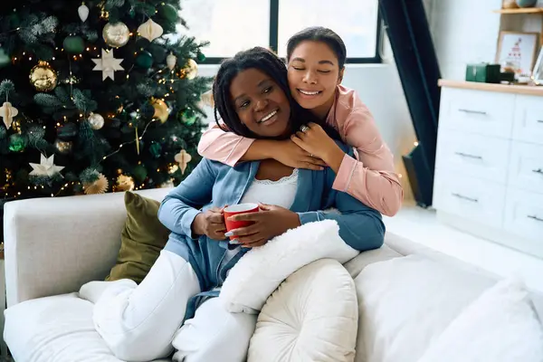 Together on a sofa, two women embrace warmly, sharing love and laughter by a festive tree. — Stock Photo