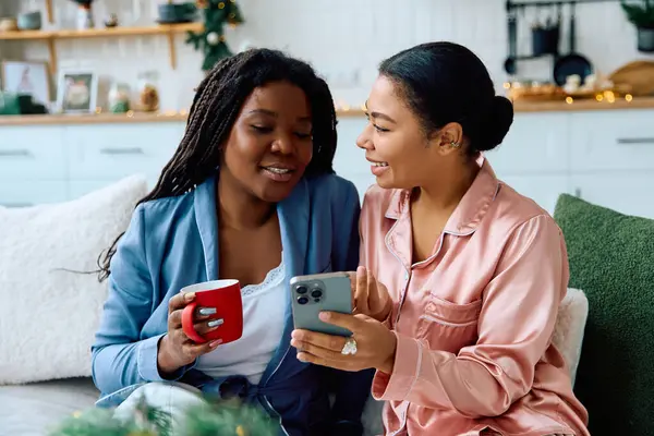 Zwei Frauen lächeln, während sie ein warmes Getränk genießen und freudige Momente teilen, während sie auf ein Telefon schauen. — Stockfoto