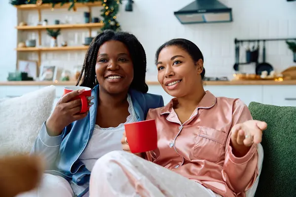 Two women in pajamas enjoy each others company while sipping hot drinks and laughing together — Stock Photo