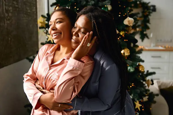 Due donne felici si abbracciano vicino a un albero di festa incandescente, condividendo un caldo momento di amore e gioia. — Foto stock