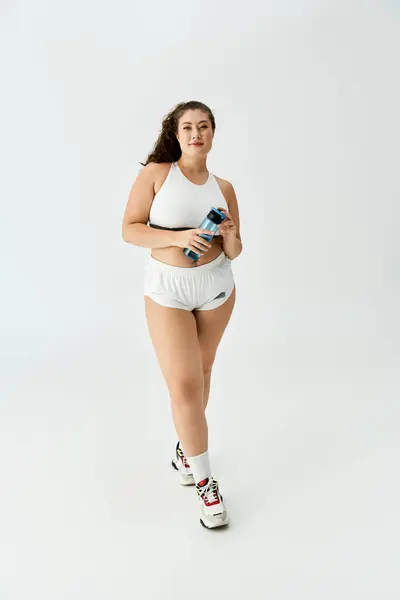 Una giovane donna plus size posa fiduciosa in activewear bianco alla moda mentre tiene in mano una bottiglia d'acqua. — Foto stock