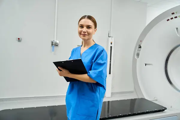 An attractive female doctor holding a clipboard stands beside an MRI machine in a hospital setting. — Foto stock