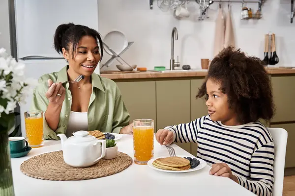 Cheerful mother and her little girl enjoy pancakes and juice together in a bright kitchen. — Stock Photo