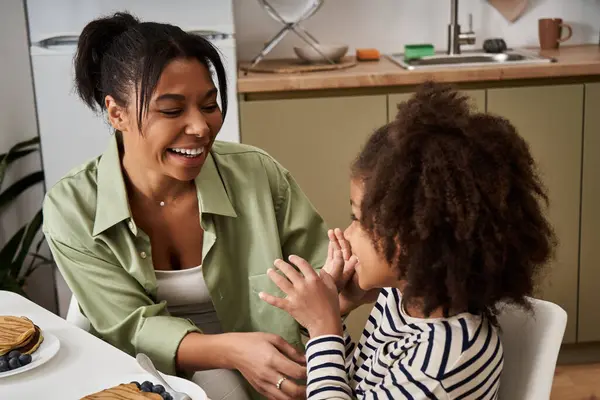 A mother smiles joyfully at her daughter while they enjoy pancakes together in their cozy kitchen. — Stock Photo