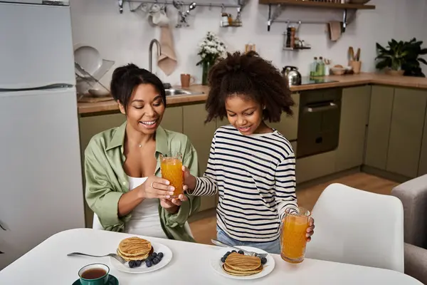 Mother and daughter joyfully toast with drinks while enjoying pancakes in a cozy kitchen. — Stock Photo