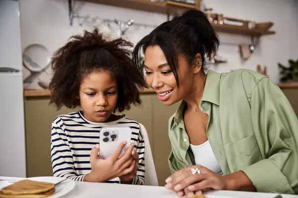 A mother and her little girl smile together as they explore something fun on a phone at home — Stock Photo