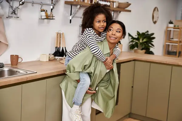 A mother carries her happy daughter on her back in a warm, stylish kitchen. — Stock Photo